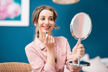 Young woman applying a lipstick sitting with a mirror in the beautiful blue bedroom at home