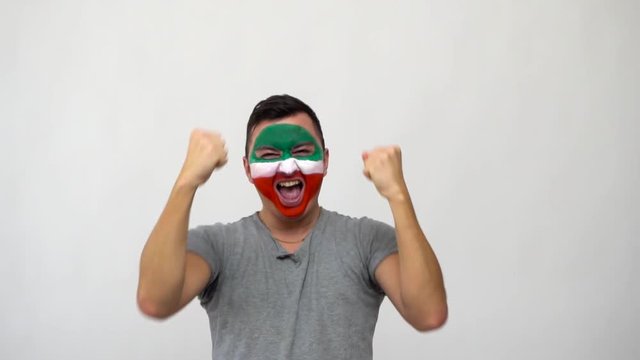 Handsome man supporter fan of Iran national team painted flag face get happy victory screaming into a camera. Football emotions.