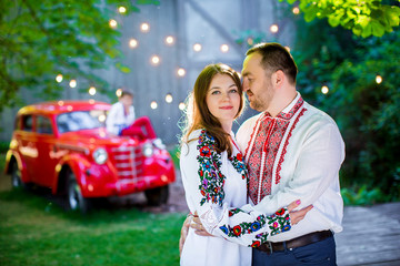 Beautiful couple in embroidery shirts , hugging on background old red car in the park