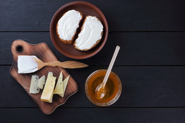 Hard cheese on a wooden board, sandwiches with soft cheese, honey in glass dishes, copy space, minimalism, cheese pattern, wooden dark background, healthy food, wooden cutlery