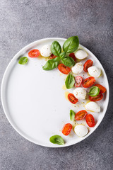 White plate of healthy classic delicious caprese salad with ripe tomatoes and mozzarella cheese with fresh basil leaves on gray concrete background with space for text. Italian food. Top view.