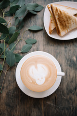 Delicious fresh morning cappuccino coffee with a funny cat face drawn on the milk foam, hot and freshly grilled club sandwich and some eucalyptus branches as a decoration, wooden table background