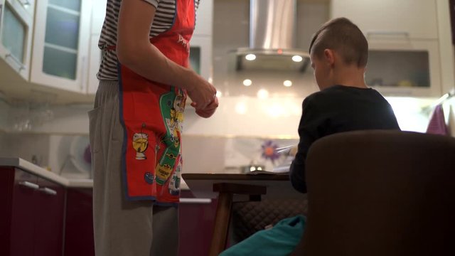 The boy flips through the pages of a book with culinary recipes. He finds the dish they cook. A father and his sons want to make a mom a surprise.