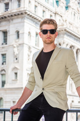 Young American Businessman Casual Fashion in New York, wearing beige blazer, black undershirt, black pants, sunglasses, sitting on railing in front of vintage office building..