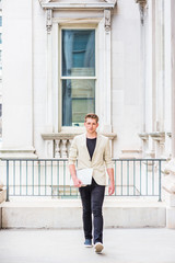 Young American Businessman with little beard working in New York, wearing beige blazer, black pants, casual shoes, holding laptop computer, walking in vintage office building, going to work..