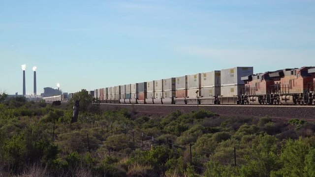 A freight train heads across the desert with an industrial site background.