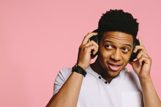 Portrait of a cool African American guy in white shirt, holding headphones and looking up, on pink background