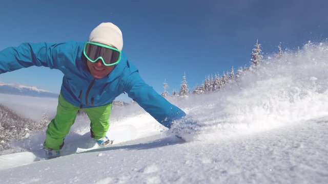 SLOW MOTION CLOSE UP PORTRAIT: Smiling snowboarder doing a cool hand drag in fresh powder snow. Stoked male snowboarder drags his hand through freshly fallen snow as he carves down the sunny mountain.