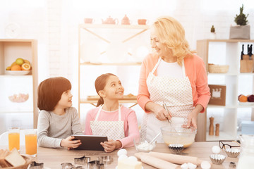 Young grandmother cooks in kitchen with her grandchildren looking at tablet.