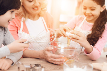 Young grandmother and grandchildren break eggs into bowl in kitchen. Llittle girl is learning to cook.