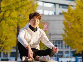 Young Chinese smiling man portrait sitting with blur yellow trees backgroundin sunset in autumn park. Outdoor.