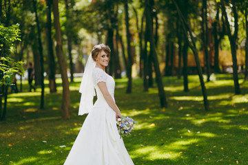 Fototapeta na wymiar Beautiful wedding photosession. Elegant young bride in white dress and veil with beautiful hairdress with bouquet of flowers and ribbons near trees on wedding walk in the big green park on sunny day