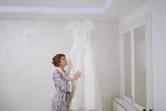 Morning preparation of the newlyweds for the wedding ceremony. The bride in a patterned robe with a beautiful hairstyle stands beside her wedding white long lace dress, which hangs on a hanger
