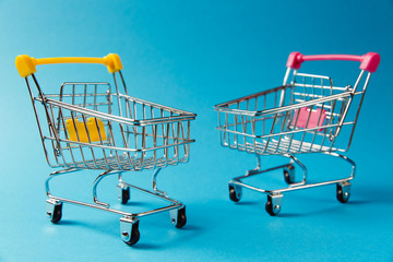 Close up of two supermarket grocery push carts for shopping with wheels and yellow and pink plastic elements on handle isolated on blue background. Concept of shopping. Copy space for advertisement