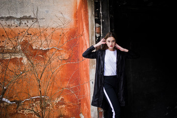 Naklejka premium Fashionable long legs brunette model in long black cloak posed outdoor at winter day against old grunge wall.