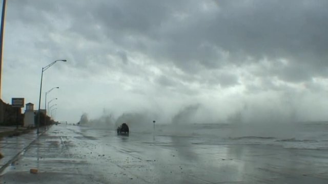 Huge waves pound a seawall in Galveston Texas during a massive hurricane or storm as a car drives by.