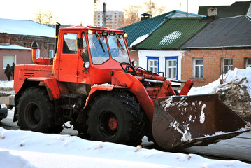 Obraz na płótnie Canvas Bright orange excavator tractor cleaning snow on the road along residential houses, snowy winter in Kharkiv, Ukraine