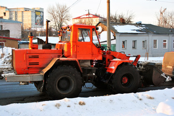 Bright orange excavator tractor cleaning snow on the road along residential houses, snowy winter in Kharkiv, Ukraine