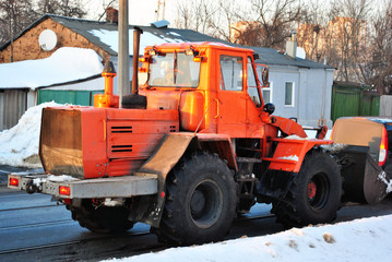 Bright orange excavator tractor cleaning snow on the road along residential houses, snowy winter in Kharkiv, Ukraine