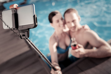 Saying cheese. Selective focus on a smartphone held by young woman and man embracing in a swimming pool while posing for a selfie together.