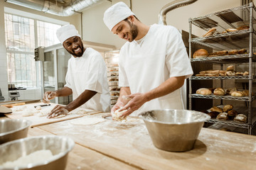 happy bakers kneading dough together at baking manufacture and talking - obrazy, fototapety, plakaty