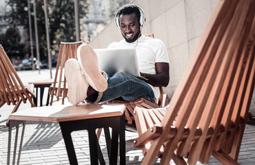 No time for worries. Relaxed young man grinning broadly while enjoying the music playing in his headphones and working on his laptop outdoors.