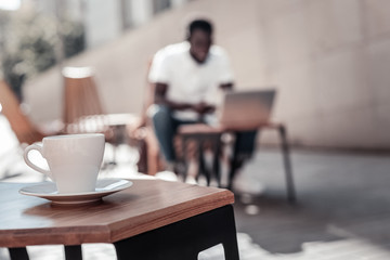 Recharge your batteries. Selective focus on a white ceramic cup of aromatic coffee standing on a wooden table with a young man browsing the Internet on his laptop over the background.