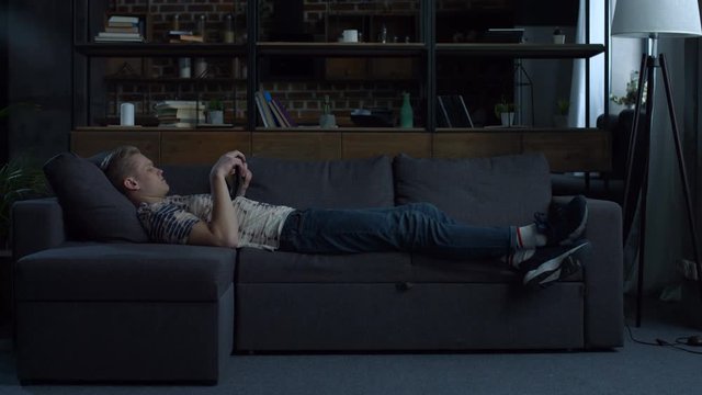 Carefree handsome young man surfing the net with digital tablet while lying on the couch at home on lazy day. Relaxed male hipster on sofa networking with tablet computer at home. Side view.