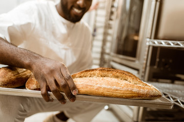 cropped shot of african american baker with tray of fresh loaves of bread on baking manufacture