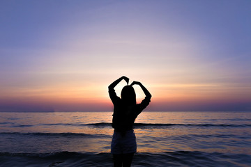 Silhouette of woman showing love, making heart shaped gestures with arm and enjoying sunset at beach