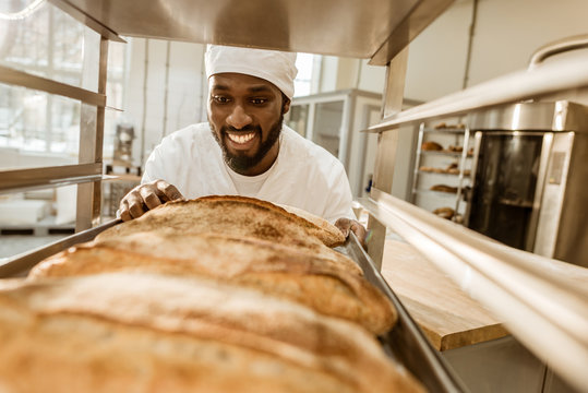 smiling african american baker looking at fresh loaves of bread on baking manufacture