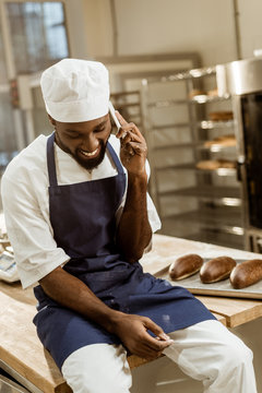 young african american baker with hands covered in flour talking by phone while sitting on work table