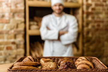 blurred baker with crossed arms standing at pastry store with basket of croissants on foreground