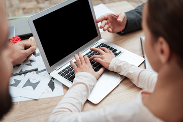 Little girl is typing on laptop that stands on table where there are sheets of graphic drawings.