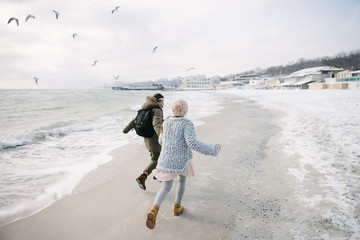 back view of young couple holding hands and running on winter seashore