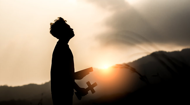 Young holding Bible book and christian cross with light sunset background