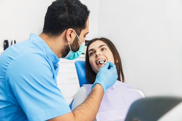 Dentist examining patient's teeth, good-looking man with black hair wearing blue uniform and gloves, looking at patient's teeth carefully with dental instruments