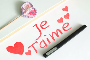 Notepad on a desktop in a workplace with the text JE T'AIME (love you) in Spanish. Some papers, a pen and an amigurumi heart.