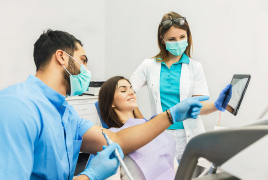 Smiling beautiful patient sitting in chair with dental purple bib, male dentist, in blue uniform and gloves, female assistant, in green mask with tablet, standing next to the patient - Powered by Adobe