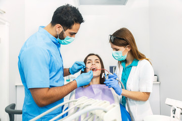 Fototapeta premium Smiling beautiful patient sitting in chair with dental purple bib, male dentist, in blue uniform and gloves, holding mouth mirror and explorer, female assistant, in green mask with saliva ejector