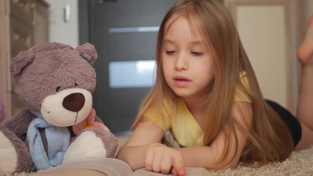 Cute happy little girl with teddy bear and reading book. Pretty kid at home, lying on the floor. Education concept