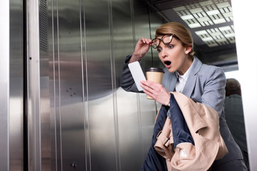 portrait of shocked businesswoman in suit with smartphone in elevator