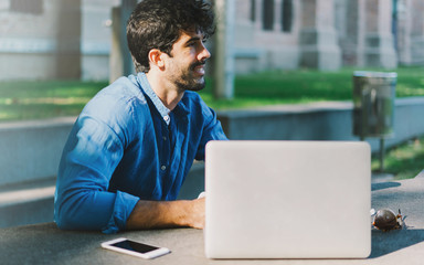 Fototapeta premium Successful freelancer thinking on a new project while sitting outdoors with a laptop and a smartphone. Hipster guy enjoying summer day in a park while sitting at the table with gadgets on it.