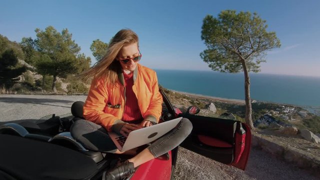 Cute millennial student or freelance designer, works remotely from office, rests on top of mountain on amazing scenery and beautiful view of ocean, types on laptop, always connected to social media