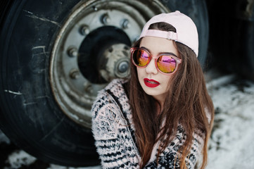 Brunette stylish casual girl in cap and sunglasses sitting against truck wheels.