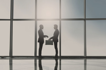 Silhouette view of two young businessmen are standing with suitcase in modern office with panoramic windows