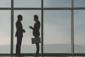 Fototapeta na wymiar Silhouette view of two young businessmen are standing in modern office with panoramic windows