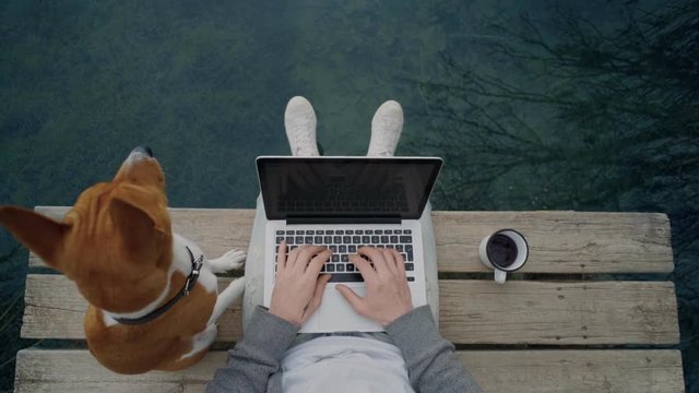 POV awesome shot of hipster man or millennial freelancer write blog post or work on remote office project on laptop, while drinking coffee from adventure metal mug and pet dog on pier on lake