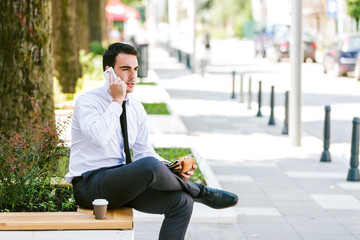 Young businessman talking on cellphone while eats sandwich and drink coffee to go