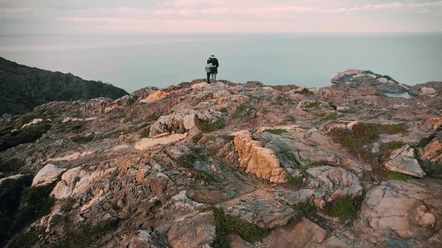 Aerial drone footage of romantic couple hug and cuddle on edge of amazing beautiful cliff or rock, overlooking mountains and ocean in inspiring and exquisite landscape, travel bloggers adventure